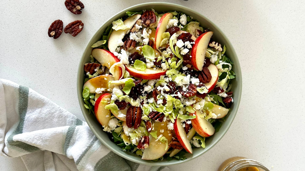 Final plated salad served on a rustic wooden table, topped with crumbled goat feta and sprinkled with dried cranberries and chopped pecans for a seasonal, nourishing fall meal