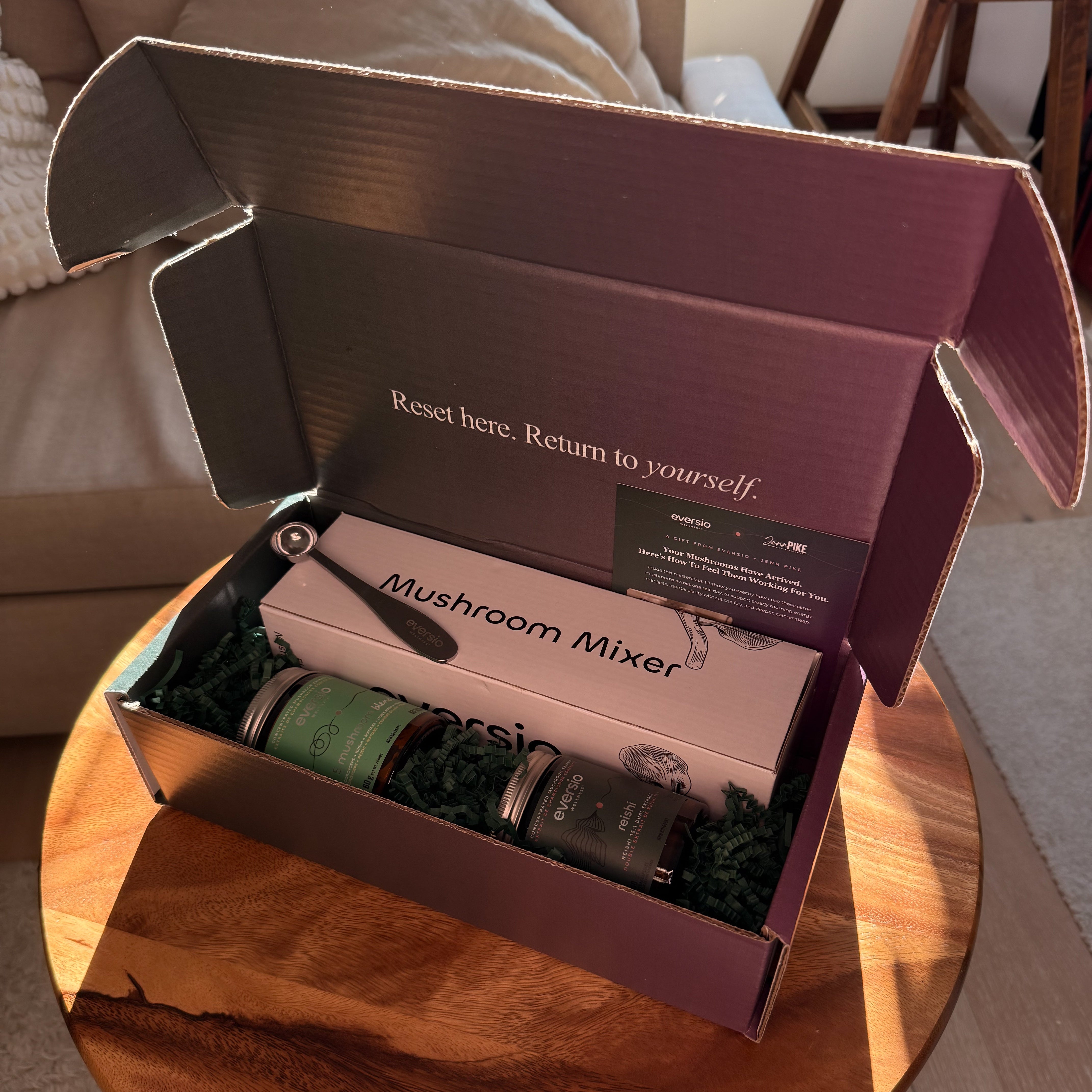 An open mauve-colored subscription box labeled 'Mushroom Mixer' sits on a round wooden table in a living room setting. Inside the box is a white box with 'Mushroom Mixer' printed on it, a small black-handled tool, and two dark glass bottles with green labels nestled in green crinkle paper.