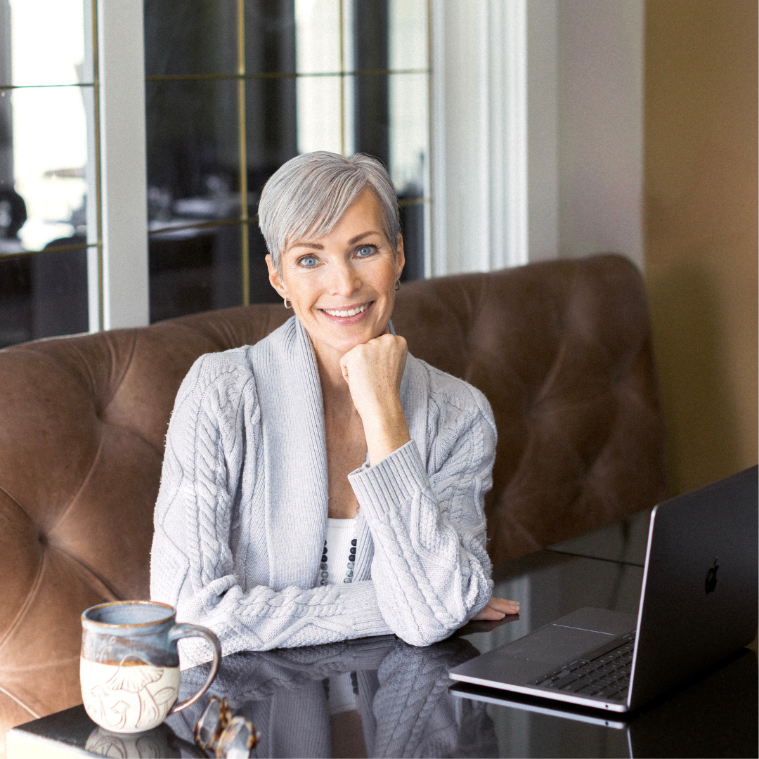 Brandi Garden sitting on a brown couch with a laptop and mushroom mug, smiling.